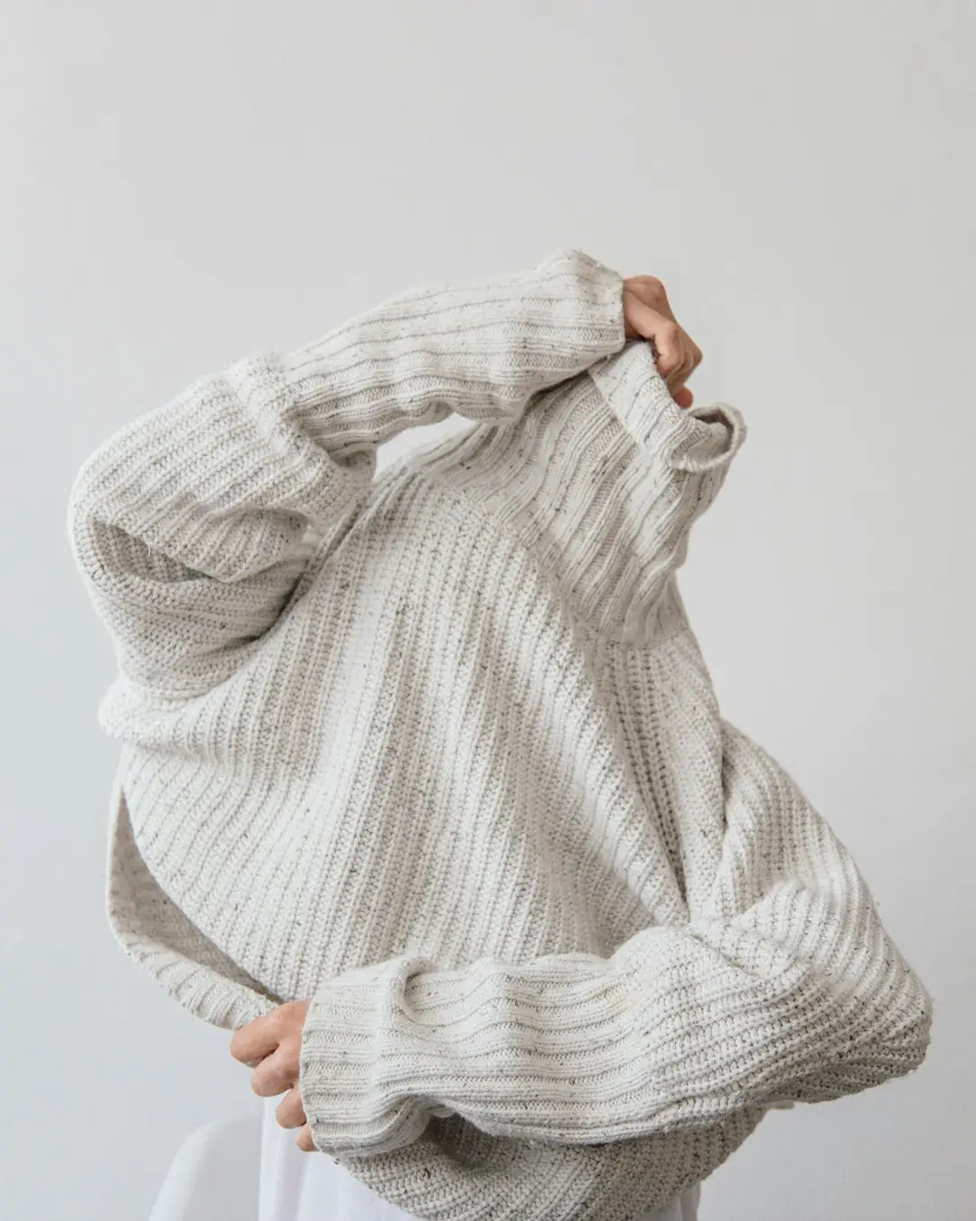 Mulher fechando um casaco de tricô graúdo sobre uma camiseta de algodão claro, mãos perto do pulso, luz suave de inverno