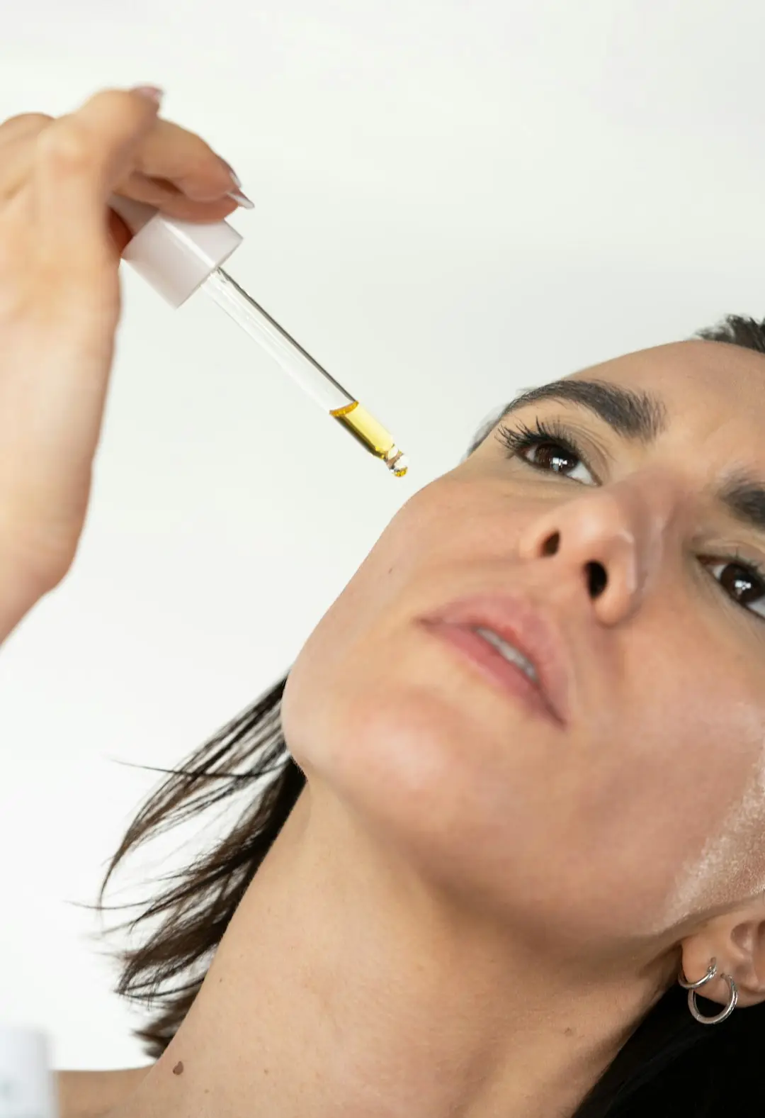 Mulher aplicando gota de sérum na bochecha diante do espelho, luz natural clara